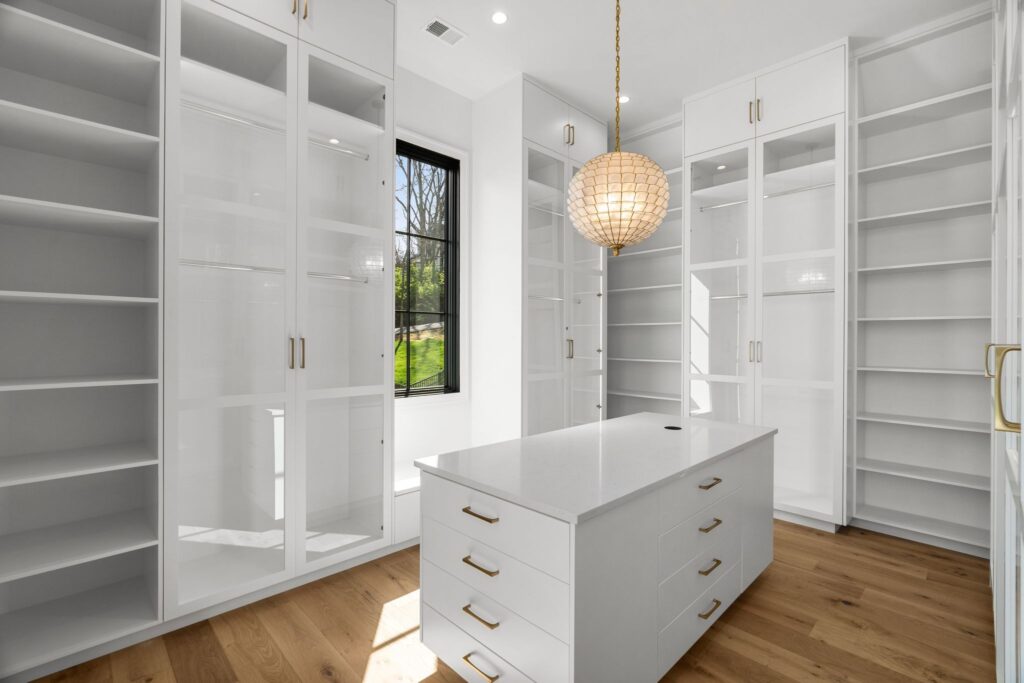 A luxurious closet space featuring a white center island, glass-fronted cabinets, and a statement light fixture by Destination Cabinets.