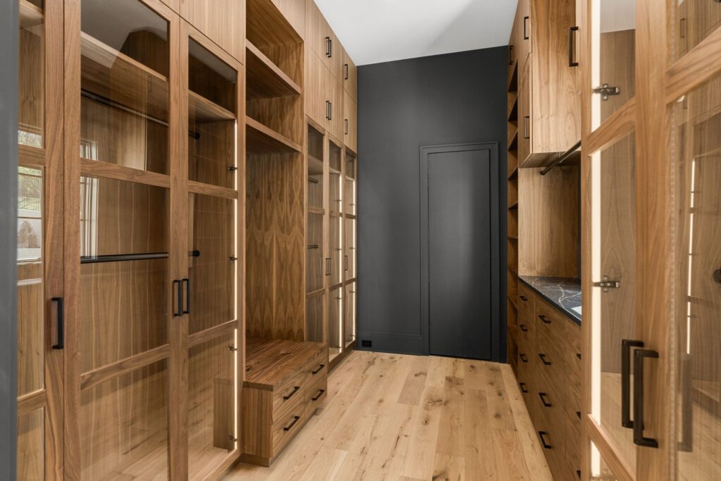 A sophisticated closet featuring natural wood cabinets with glass fronts, a striking black backdrop, and light wood flooring by Destination Cabinets.