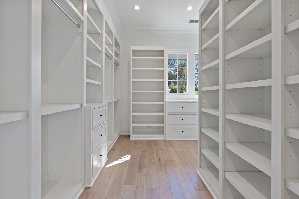 A bright closet with pure white shelving and cabinetry complemented by natural light and light wooden flooring by Destination Cabinets.