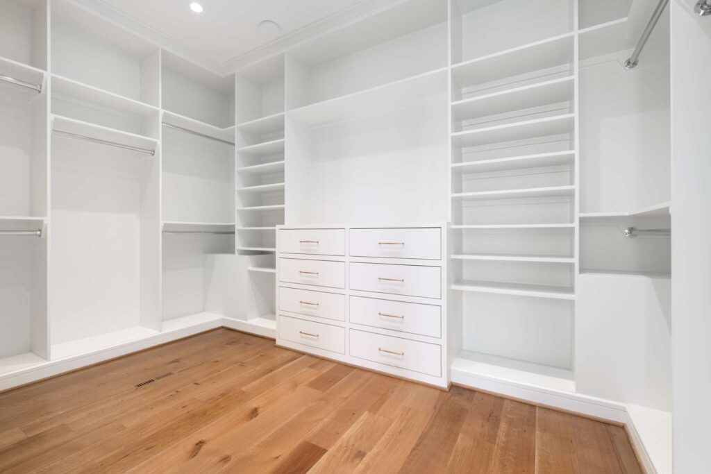 A bright and airy closet featuring white custom cabinetry and drawers against a warm wooden floor by Destination Cabinets.