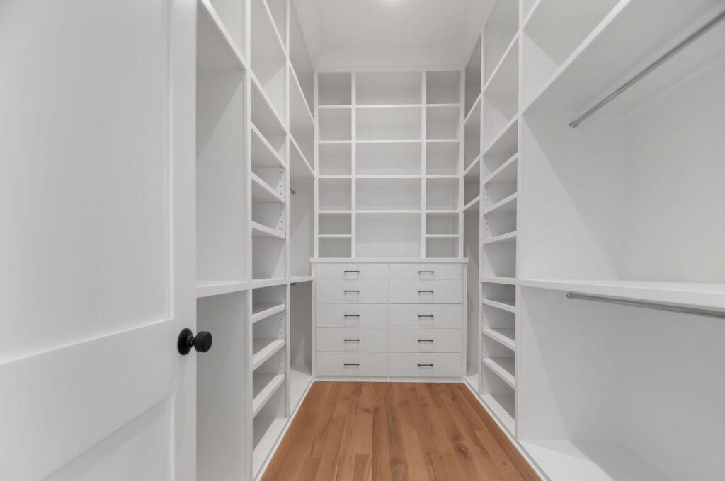 A clean and bright closet with white custom drawer units and shelving, accented by warm wooden flooring by Destination Cabinets.