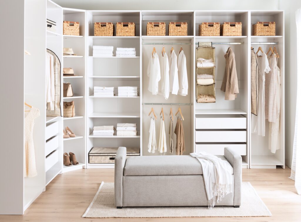 A well-organized closet with white shelving, woven baskets, and a neutral-toned seating area by Destination Cabinets.