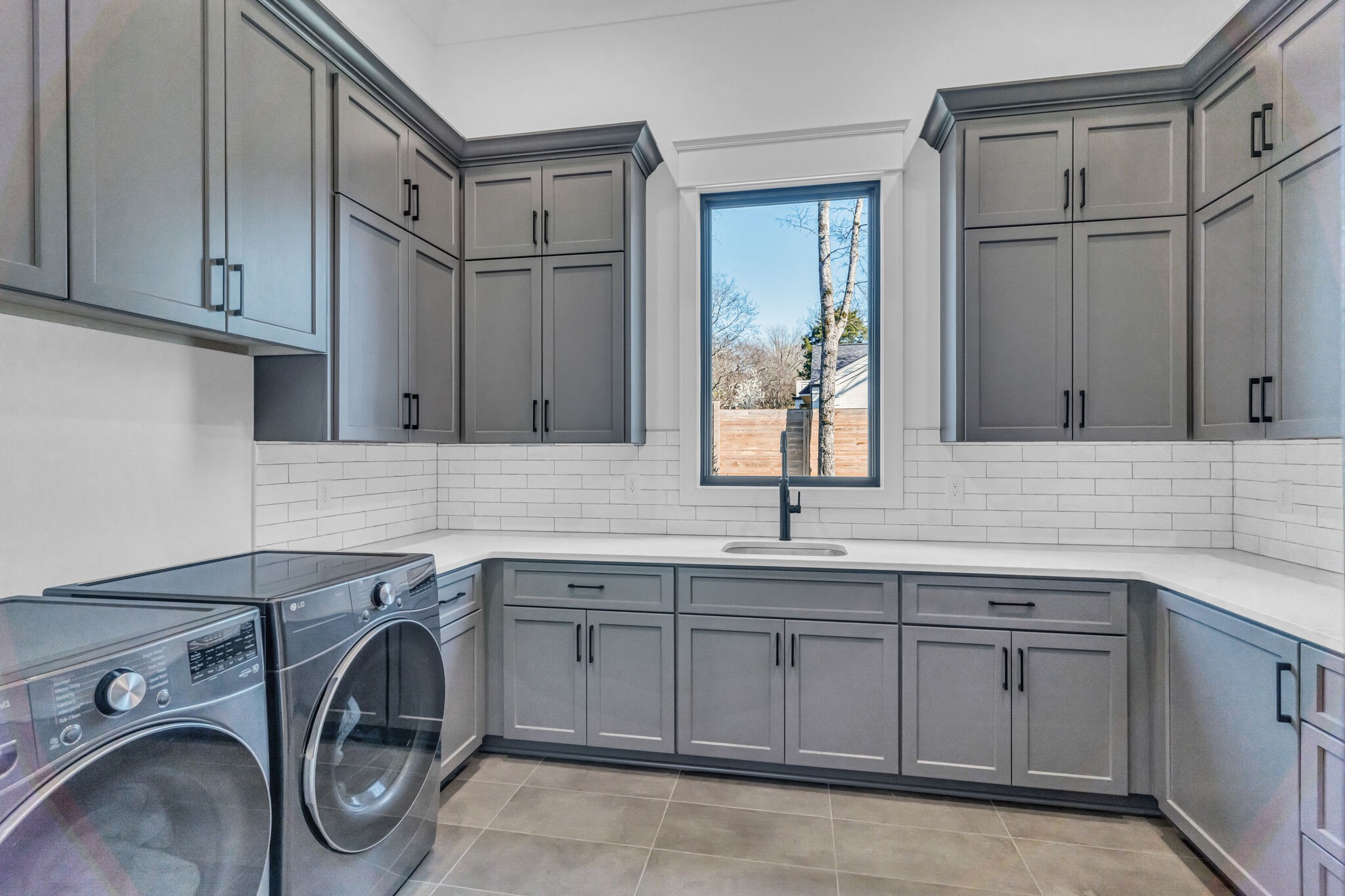 Spacious laundry room featuring gray cabinets with contemporary hardware, subway tile backsplash, and advanced washing appliances, by Destination Cabinets.